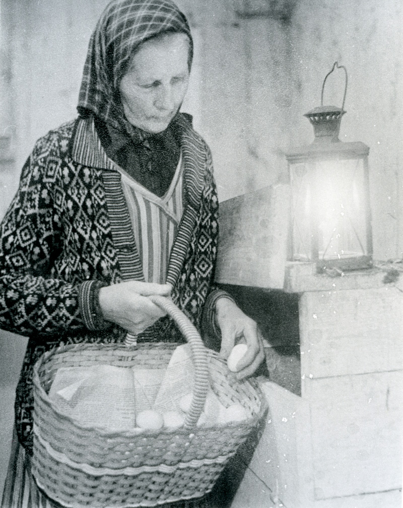 Svartvitt fotografi. Kvinna med huckle plockar ägg från en hylla ned i en korg.