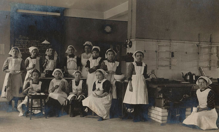 Flickor med vita förkläden uppställda framför kameran i ett kök.