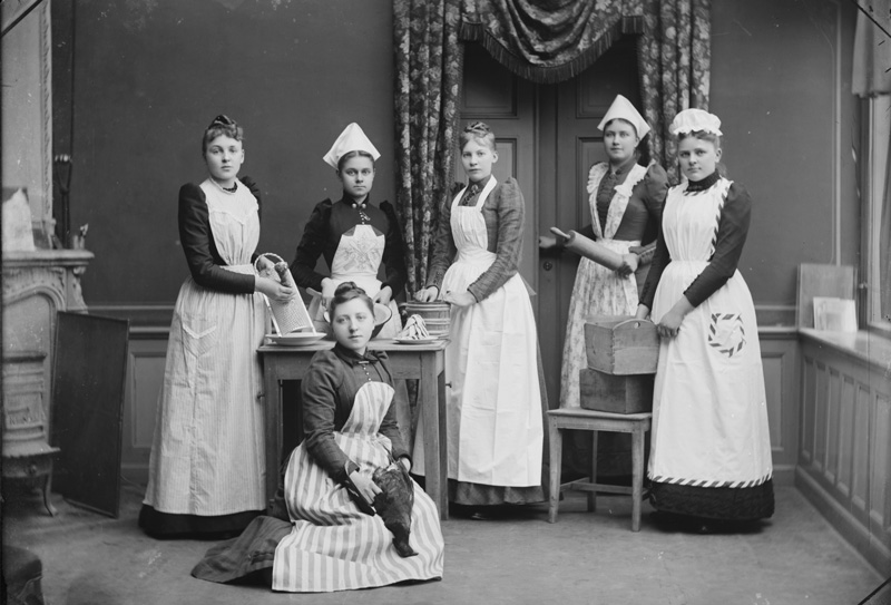 Svartvitt fotografi. Gruppbild av sex kvinnor i vita förkläden och med olika hushållsföremål som kavel och rivjärn i händerna. Alla tittar in i kameran.