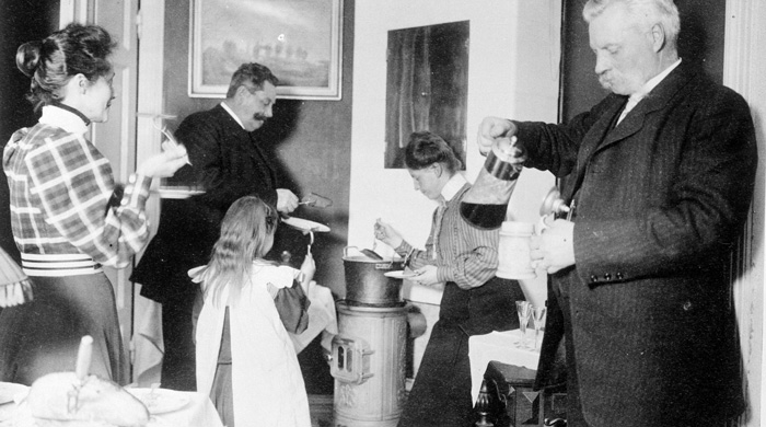 svartvitt fotografi med en familj som doppar i bröd i gryta med skinkspad som står på järnkamin. I förgrunden häller äldre man upp mumma i ett stop.