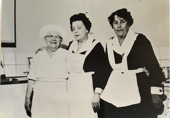 Svartvitt fotografi föreställande tre kvinnor i ett kök. Kvinnan längst till vänster har vita kockkläder på sig medan de andra kvinnorna har serveringskläder - mörka klänningar och vita förkläden.