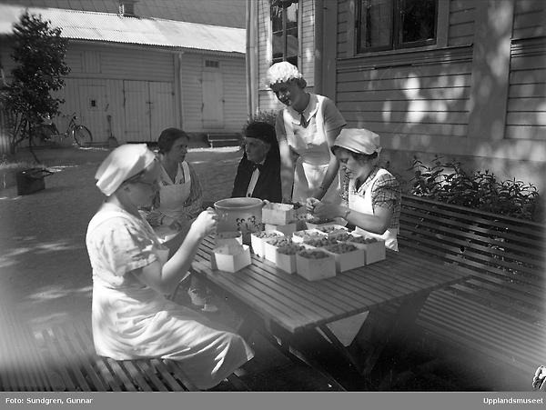 svartvitt fotografi av vitklädda kvinnor som sitter runt bord och rensar hallon