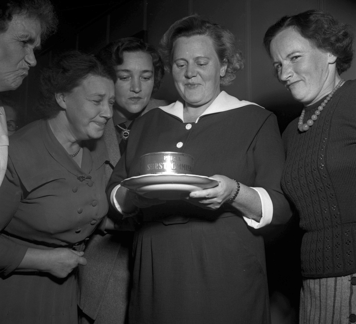 Svartvitt fotografi. Kvinna håller ett fat med en surströmmingsburk och runtomkring henne står fyra andra kvinnor, ett par av dem grimaserar över lukten.