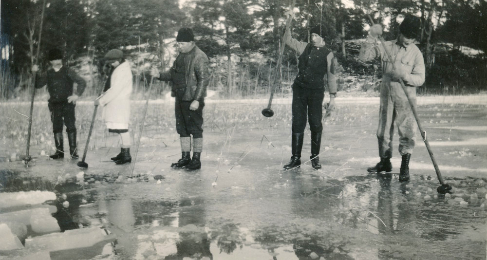 Svartviutt fotografi. Fem personer står på isen i en vik och stöter i vattnet med långa störar.