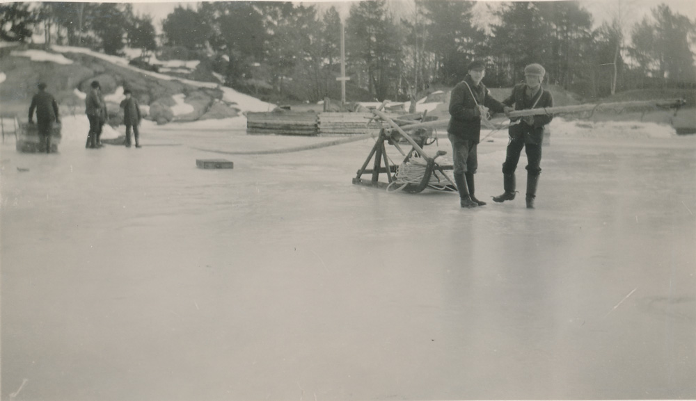 Svartvitt fotografi. Två män som drar eb träställning över isen. I bakgrunden syns flera personer.