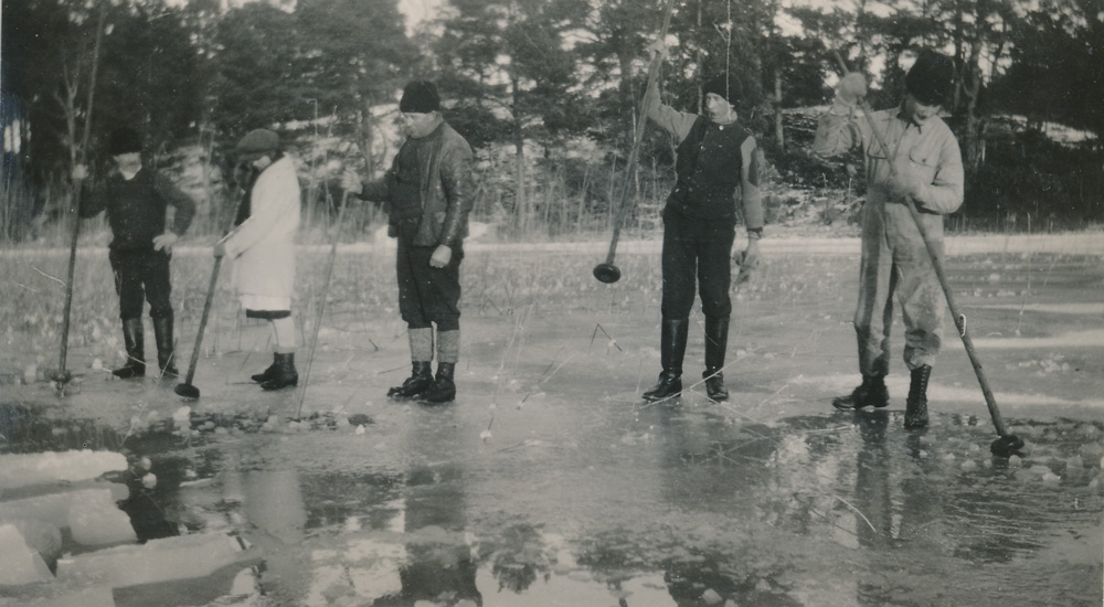 Svartvitt fotografi föreställande flera män som står bredvid varandra på isen och stöter med stora störar i vattnet. 