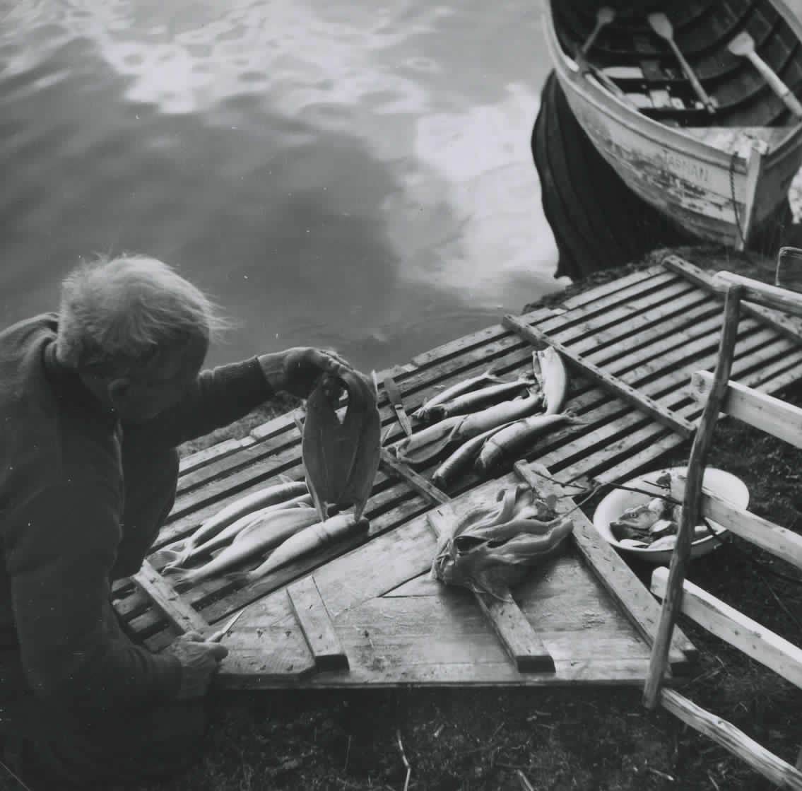 Svartvitt fotografi. En man sitter på huk vid brygga och rensar fisk.