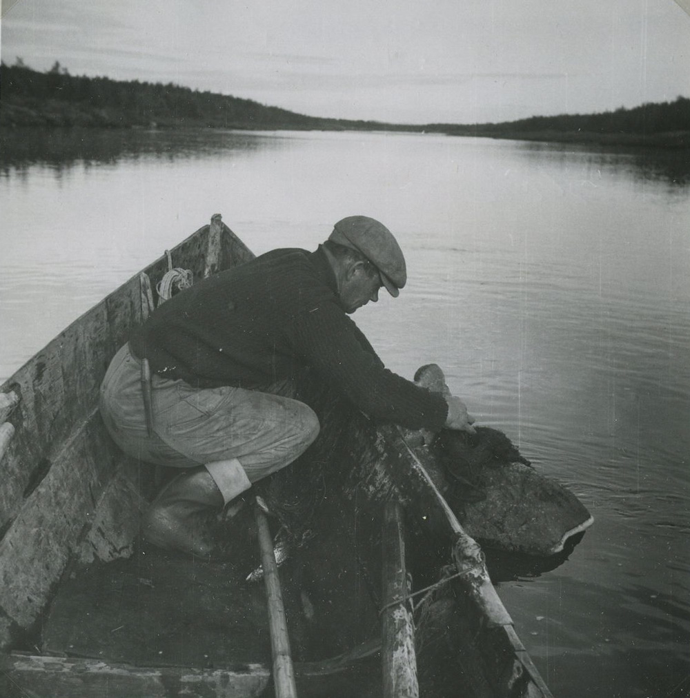 Svartvitt fotografi. En man sitter i fören på en båt och drar in nät från vattnet.